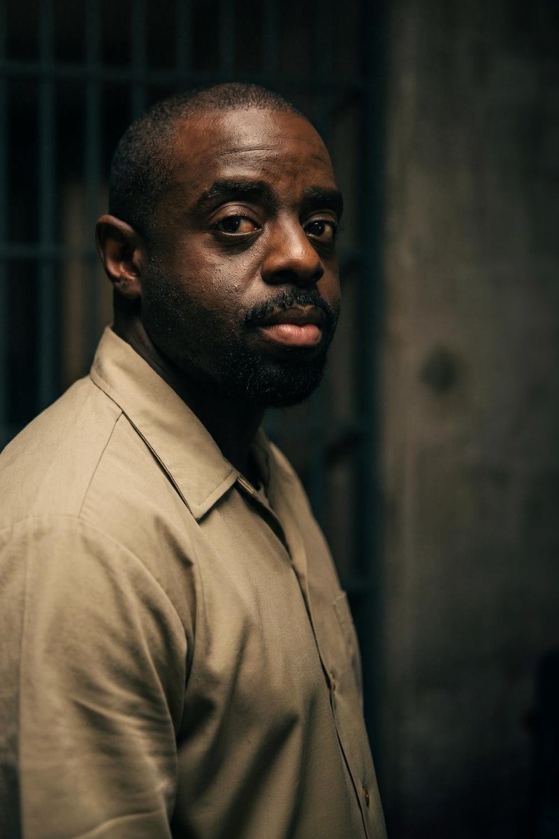 Akeem Mair as Tee, bearded and watchful, in a beige prison shirt